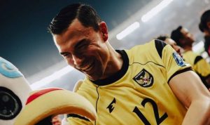 Seorang pemain tim nasional sepak bola Indonesia mengenakan jersey kuning dengan logo Garuda, tersenyum lebar dan gembira di samping maskot atau figur lain di lapangan.
