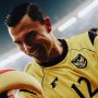 Seorang pemain tim nasional sepak bola Indonesia mengenakan jersey kuning dengan logo Garuda, tersenyum lebar dan gembira di samping maskot atau figur lain di lapangan.
