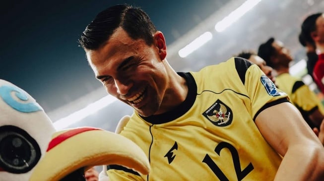 Seorang pemain tim nasional sepak bola Indonesia mengenakan jersey kuning dengan logo Garuda, tersenyum lebar dan gembira di samping maskot atau figur lain di lapangan.