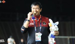 Pelatih Indra Sjafri tersenyum gembira sambil menggigit medali emas dan memegang boneka maskot kelinci, mengenakan jaket tim nasional Indonesia di stadion