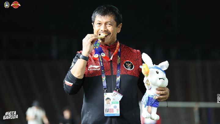 Pelatih Indra Sjafri tersenyum gembira sambil menggigit medali emas dan memegang boneka maskot kelinci, mengenakan jaket tim nasional Indonesia di stadion