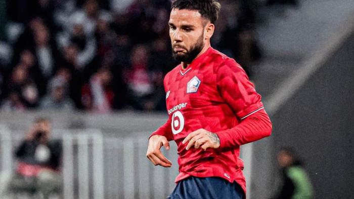 Seorang pemain Lille, Calvin Verdonk, dengan jersey merah dan celana biru tua berjalan di lapangan sepak bola, dengan ekspresi fokus. Penonton terlihat samar di latar belakang stadion.