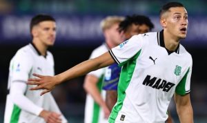 Seorang pemain Sassuolo mengenakan jersey putih-hijau dengan lengan terentang dan ekspresi fokus di tengah pertandingan sepak bola, dengan rekan setim dan pemain lawan di latar belakang.