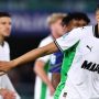 Seorang pemain Sassuolo mengenakan jersey putih-hijau dengan lengan terentang dan ekspresi fokus di tengah pertandingan sepak bola, dengan rekan setim dan pemain lawan di latar belakang.