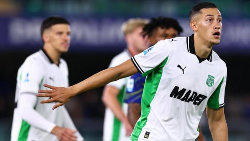 Seorang pemain Sassuolo mengenakan jersey putih-hijau dengan lengan terentang dan ekspresi fokus di tengah pertandingan sepak bola, dengan rekan setim dan pemain lawan di latar belakang.
