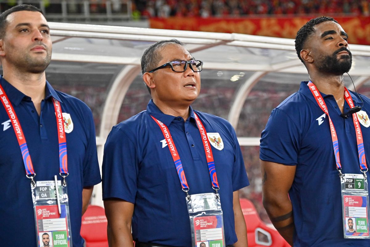 Tiga staf atau ofisial tim nasional sepak bola Indonesia mengenakan kaos polo biru gelap dengan logo Garuda, berdiri di tepi lapangan, terlihat fokus mengamati pertandingan atau persiapan tim.