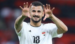 Aymen Hussein pemain sepak bola Timnas Irak dengan jersey putih dan nomor punggung 18, tersenyum dan mengangkat kedua tangannya seolah menyapa atau merayakan