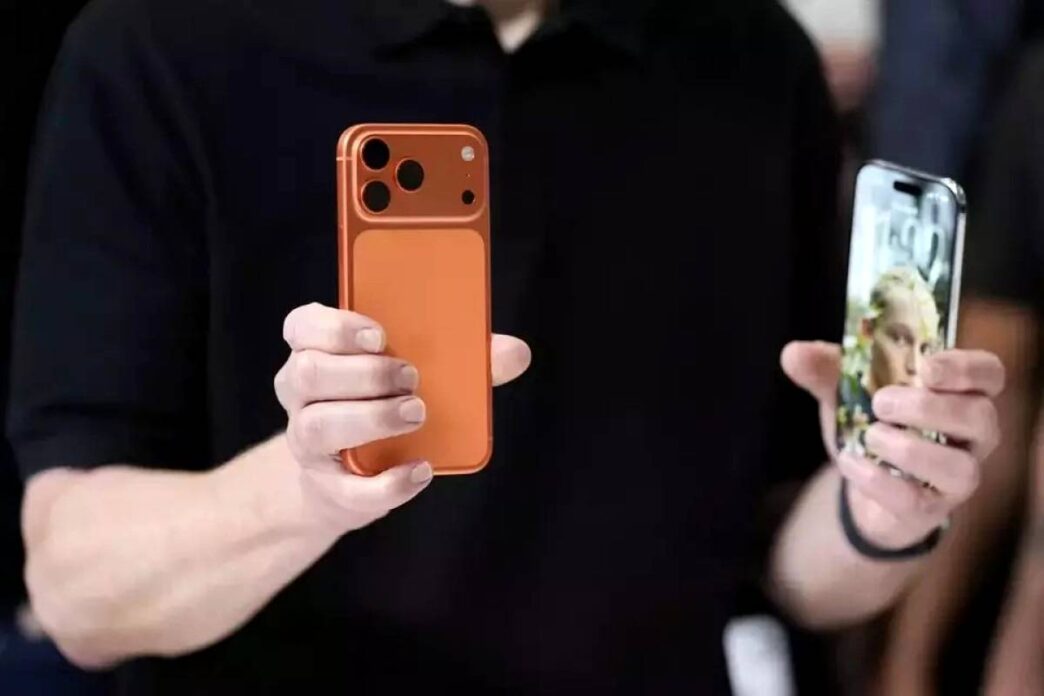 iPhone-17-Penjualan-Fantastis-Model-Baru-Warna-Oranye.webp Dua tangan memegang dua model iPhone terbaru, satu model berwarna oranye dari belakang dan satu model dengan layar menyala menampilkan wajah seorang wanita.