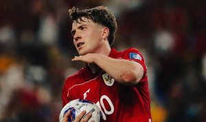 pemain tim nasional sepak bola Indonesia, Ole Romeny berambut cokelat muda, mengenakan jersey merah dengan logo Garuda dan nomor punggung 10, sedang memegang bola di lapangan dengan ekspresi serius dan fokus.
