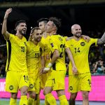 Perayaan-Pemain-Sepakbola-Malaysia-Ancaman-FIFA.jpg Sekelompok pemain sepak bola Malaysia (atau tim berseragam kuning) merayakan gol dengan ekspresi gembira dan tangan terangkat di hadapan tribun penonton yang penuh.