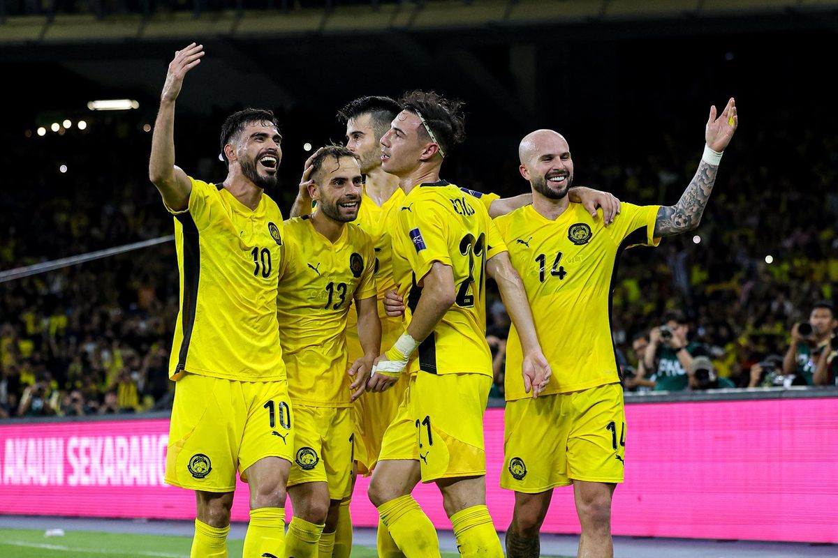 Sekelompok pemain sepak bola Malaysia (atau tim berseragam kuning) merayakan gol dengan ekspresi gembira dan tangan terangkat di hadapan tribun penonton yang penuh.