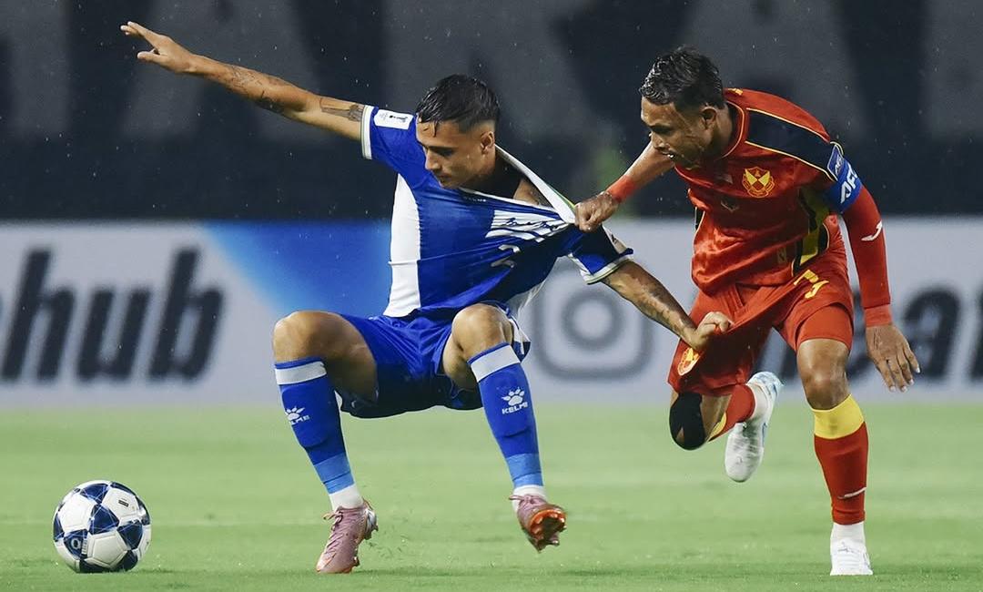 Dua pemain sepak bola, satu berseragam biru-putih dan satu berseragam merah-kuning, berduel di lapangan, menunjukkan intensitas pertandingan.