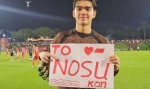 mike-rajasa-kiper-fc-utrecht-mamasa-debut-sensasional.jpeg Mike Rajasa, kiper Timnas Indonesia U-17 (jersey cokelat), tersenyum sambil memegang papan bertuliskan 'TO NOSU kan' di lapangan stadion.