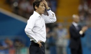 Pelatih sepak bola pria Asia (berkemeja putih) di tepi lapangan, memegang kepala dengan ekspresi tegang atau frustrasi. Timnas Indonesia