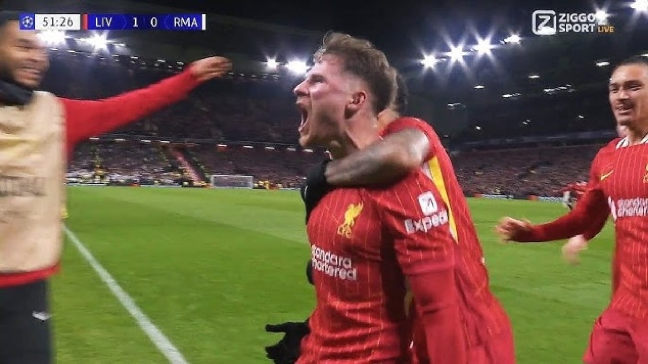 Pemain Liverpool berseragam merah merayakan gol dengan teriakan kegembiraan, memeluk rekan setim di lapangan, menunjukkan skor "LIV 1-0 RMA" di papan skor.