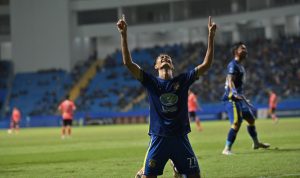 Pemain sepak bola (kostum biru) sedang merayakan gol atau menunjukkan ekspresi penuh semangat di lapangan, namun dengan nuansa ketegangan.