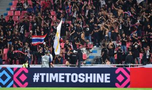 Suporter Thailand bersorak gembira di tribun stadion, beberapa membawa bendera Thailand, dengan spanduk "#TIMETOSHINE" di depan.