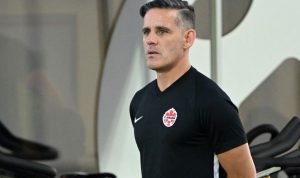 John Herdman mengenakan seragam pelatih hitam dengan logo sepak bola Kanada, memberikan instruksi di lapangan.
