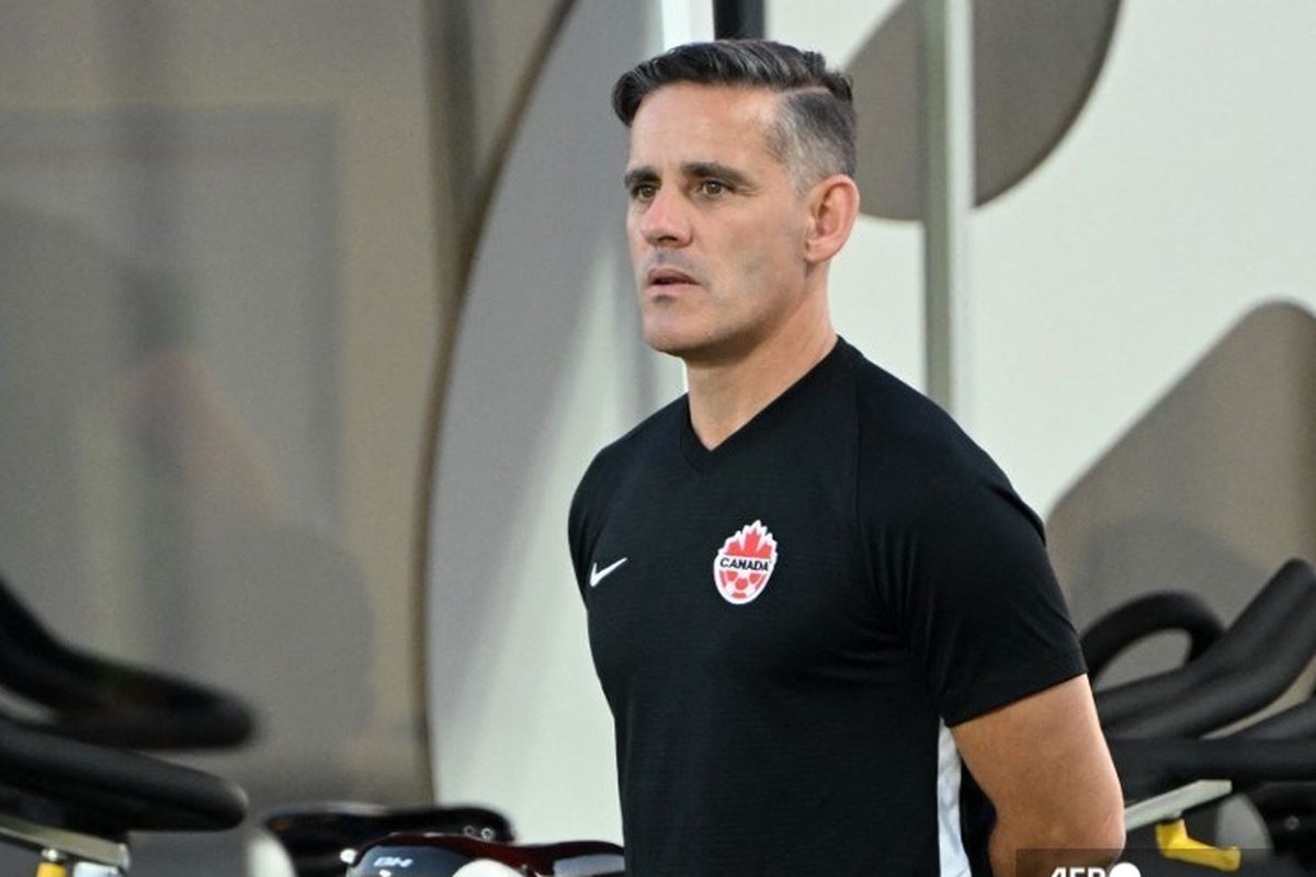 John Herdman mengenakan seragam pelatih hitam dengan logo sepak bola Kanada, memberikan instruksi di lapangan.