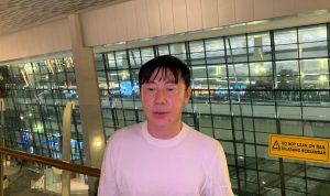 Pria paruh baya berkaos putih, (kemungkinan pengamat atau agen terkait) berdiri di area publik seperti bandara atau mal, membahas potensi Shin Tae-yong melatih Timnas.