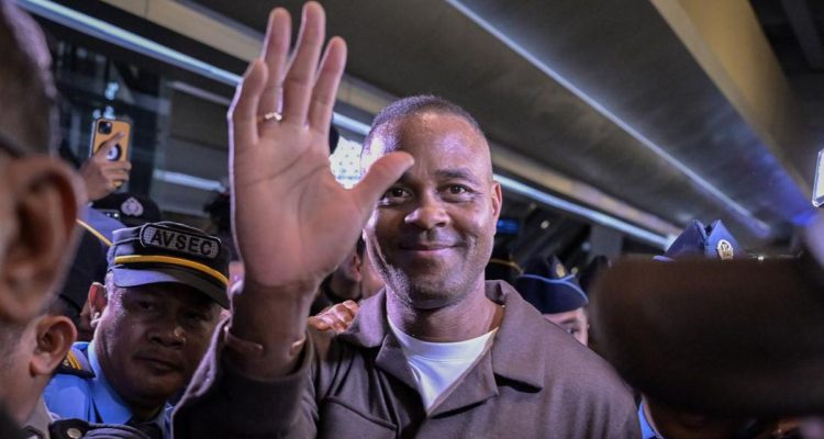 Legenda sepak bola Patrick Kluivert tersenyum dan melambaikan tangan saat tiba di bandara Indonesia dengan pengawalan petugas keamanan.