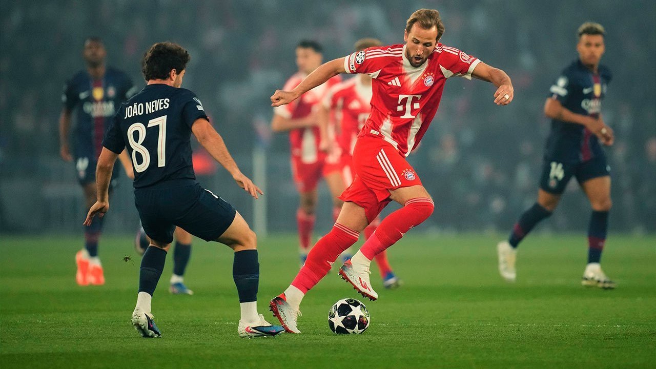 Harry Kane dari Bayern München sedang mendribel bola melewati pemain PSG João Neves dalam pertandingan Liga Champions di Allianz Arena.