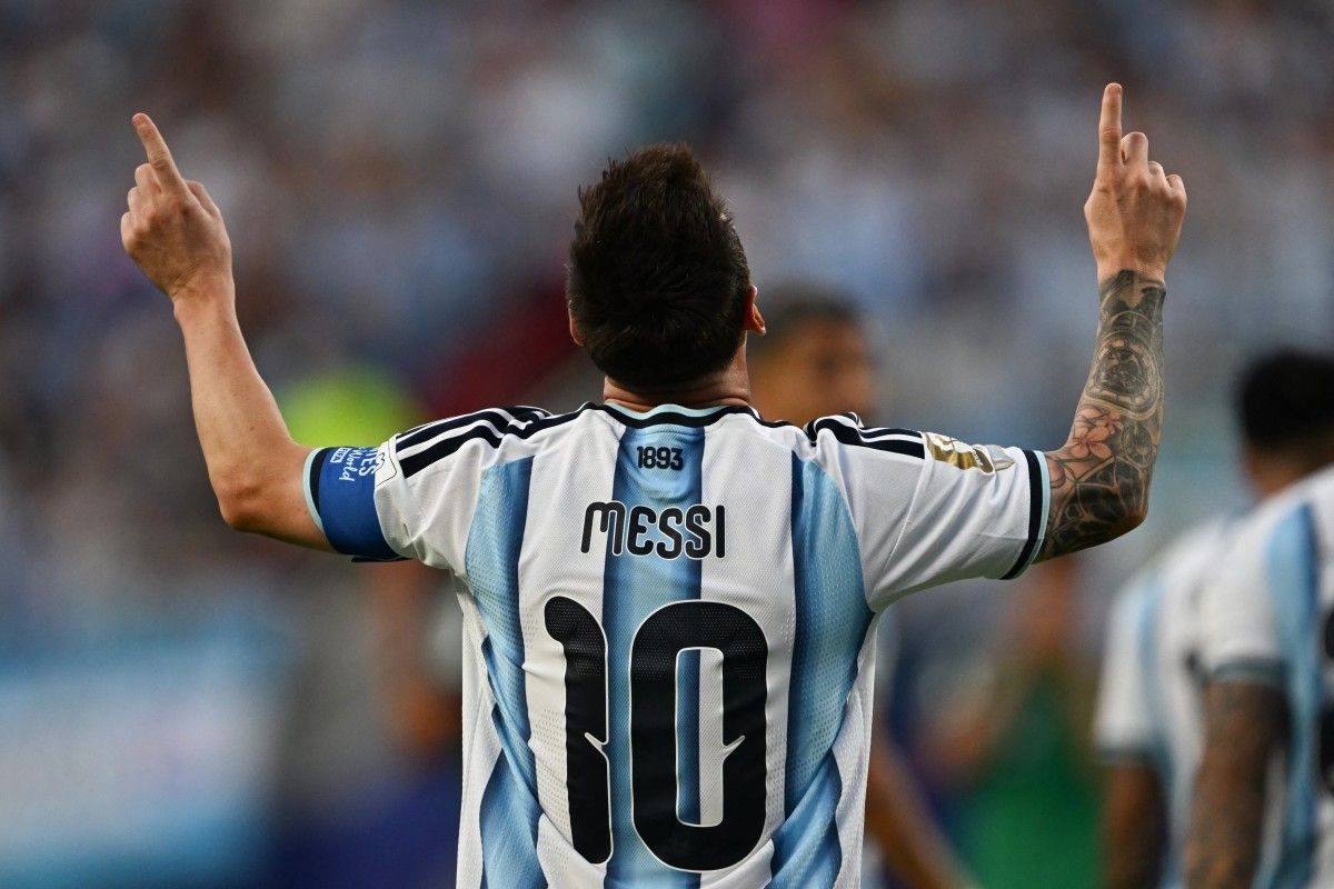 Tampak belakang Lionel Messi mengenakan jersey timnas Argentina nomor punggung 10 sambil mengangkat kedua jari telunjuknya ke langit dalam selebrasi ikonisnya.