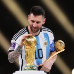 lionel-messi-pensiun-setelah-piala-dunia-2026.webp Lionel Messi tersenyum sambil memegang trofi Piala Dunia dan penghargaan Golden Ball mengenakan jersey timnas Argentina.