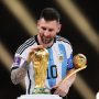 Lionel Messi tersenyum sambil memegang trofi Piala Dunia dan penghargaan Golden Ball mengenakan jersey timnas Argentina.
