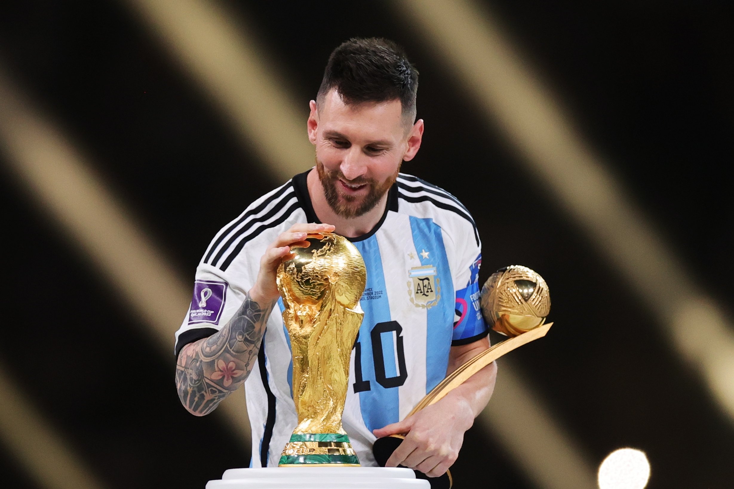 Lionel Messi tersenyum sambil memegang trofi Piala Dunia dan penghargaan Golden Ball mengenakan jersey timnas Argentina.