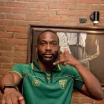 Yann Kevin Mabella mengenakan jersey hijau Persebaya Surabaya berpose dengan gestur telepon dengan latar belakang dinding bata.