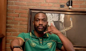 Yann Kevin Mabella mengenakan jersey hijau Persebaya Surabaya berpose dengan gestur telepon dengan latar belakang dinding bata.