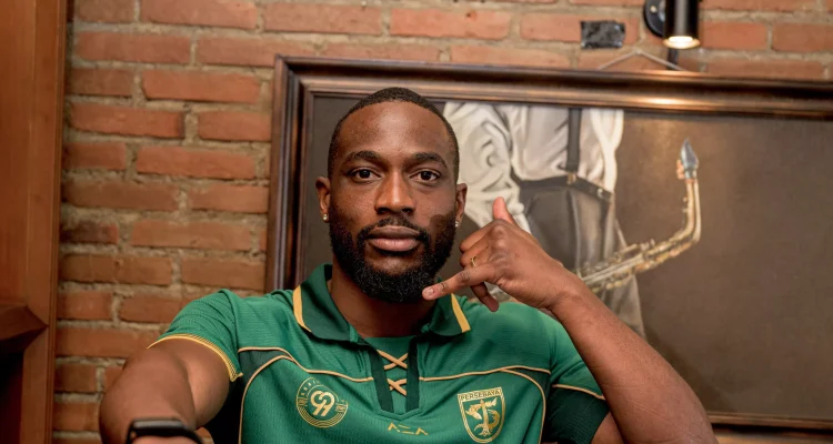 Yann Kevin Mabella mengenakan jersey hijau Persebaya Surabaya berpose dengan gestur telepon dengan latar belakang dinding bata.