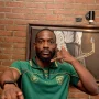 Yann Kevin Mabella mengenakan jersey hijau Persebaya Surabaya berpose dengan gestur telepon dengan latar belakang dinding bata.