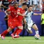 Lionel Messi dengan seragam timnas Argentina sedang menggiring bola dengan lincah melewati hadangan dua pemain lawan di lapangan hijau.
