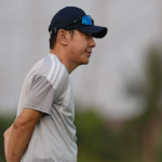 Pelatih Shin Tae-yong tampak dari samping mengenakan topi hitam dan kaus abu-abu sedang memantau di lapangan. Persija Jakarta