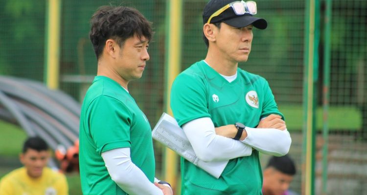shin-tae-yong-bersama-asisten-pelatih-timnas.webp Pelatih Shin Tae-yong mengenakan topi hitam dan baju hijau sedang bersedekap mengamati sesi latihan Timnas.