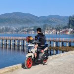 Pengendara sedang memarkir motor Yamaha GEAR ULTIMA dengan latar belakang pemandangan danau dan pegunungan.