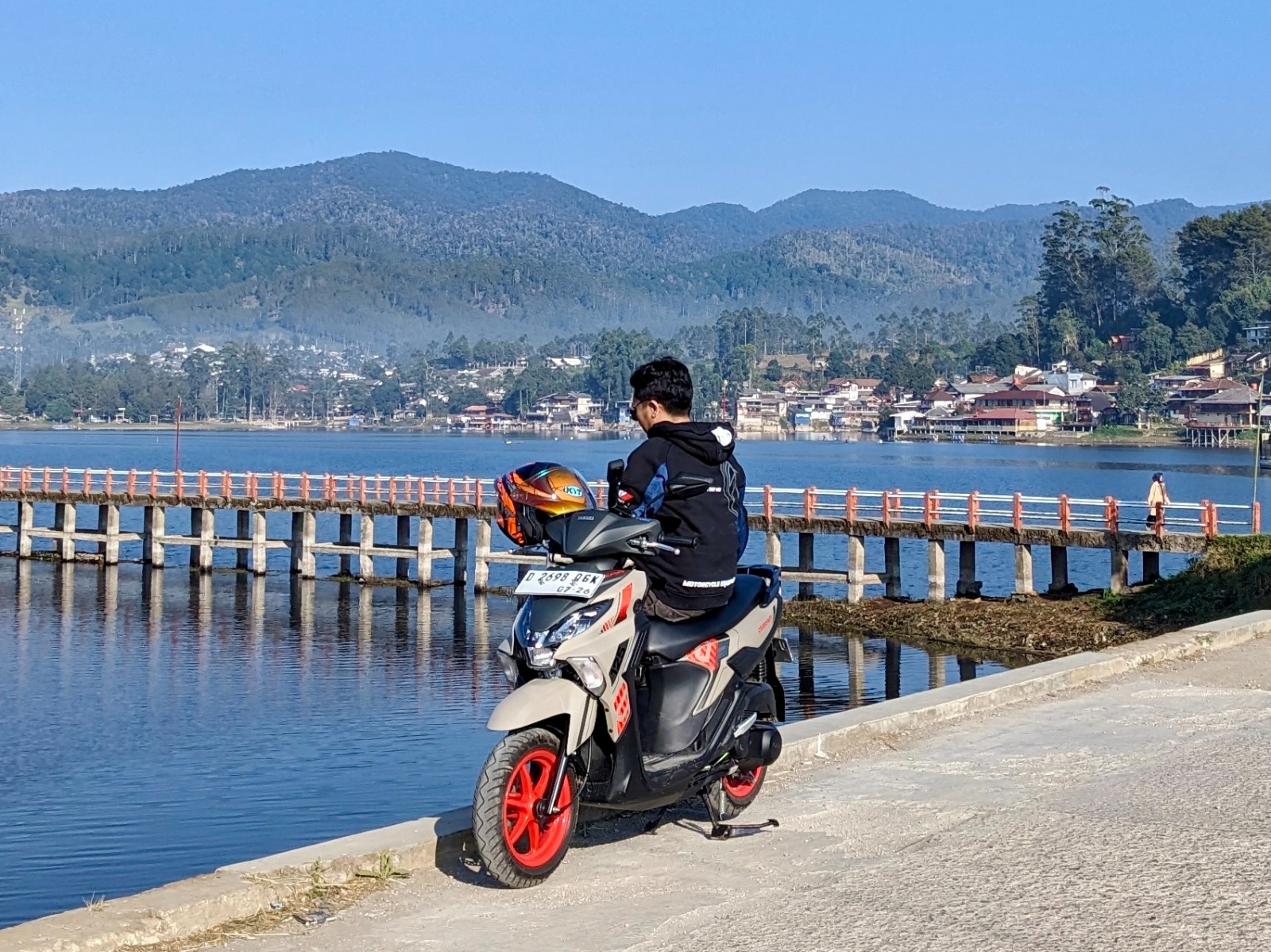 Pengendara sedang memarkir motor Yamaha GEAR ULTIMA dengan latar belakang pemandangan danau dan pegunungan.