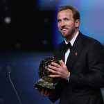Harry Kane tersenyum bangga sambil memegang trofi Ballon d'Or 2026 di atas panggung penghargaan.